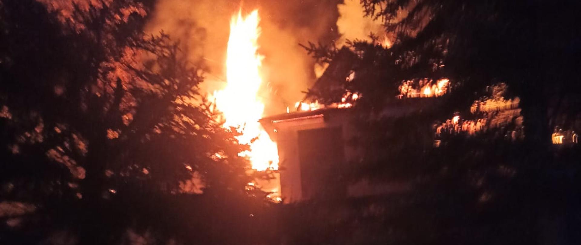 Na zdjęciu widać palący się dach budynku mieszkalnego znajdującego się między drzewami. W dole zdjęcia widać strażaka ubranego w sprzęt ochrony układu oddechowego idącego w kierunku płonącego obiektu.