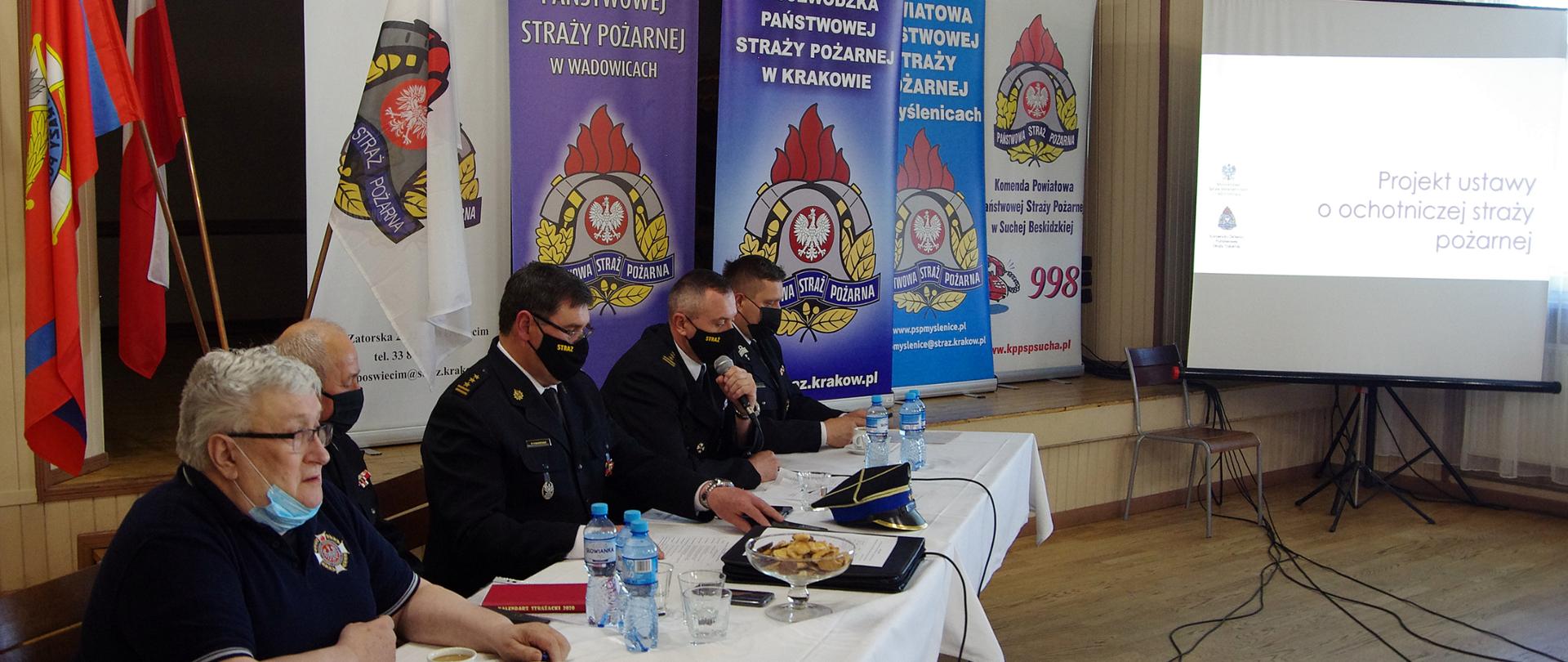 Widok z półprofilu. Na pierwszym widoczny stół prezydialny, za którym siedzą w mundurach strażackich i maseczkach ochronnych oficerowie PSP i członkowie OSP. W centralnej części siedzi Małopolski Komendant Wojewódzki PSP i omawia tekst projektu ustawy o OSP. W tle za stołem prezydialnym widoczne banery KW PSP Kraków i kilku KP PSP. Po prawej stronie ekran z tekstem projektu ustawy o PSP. 