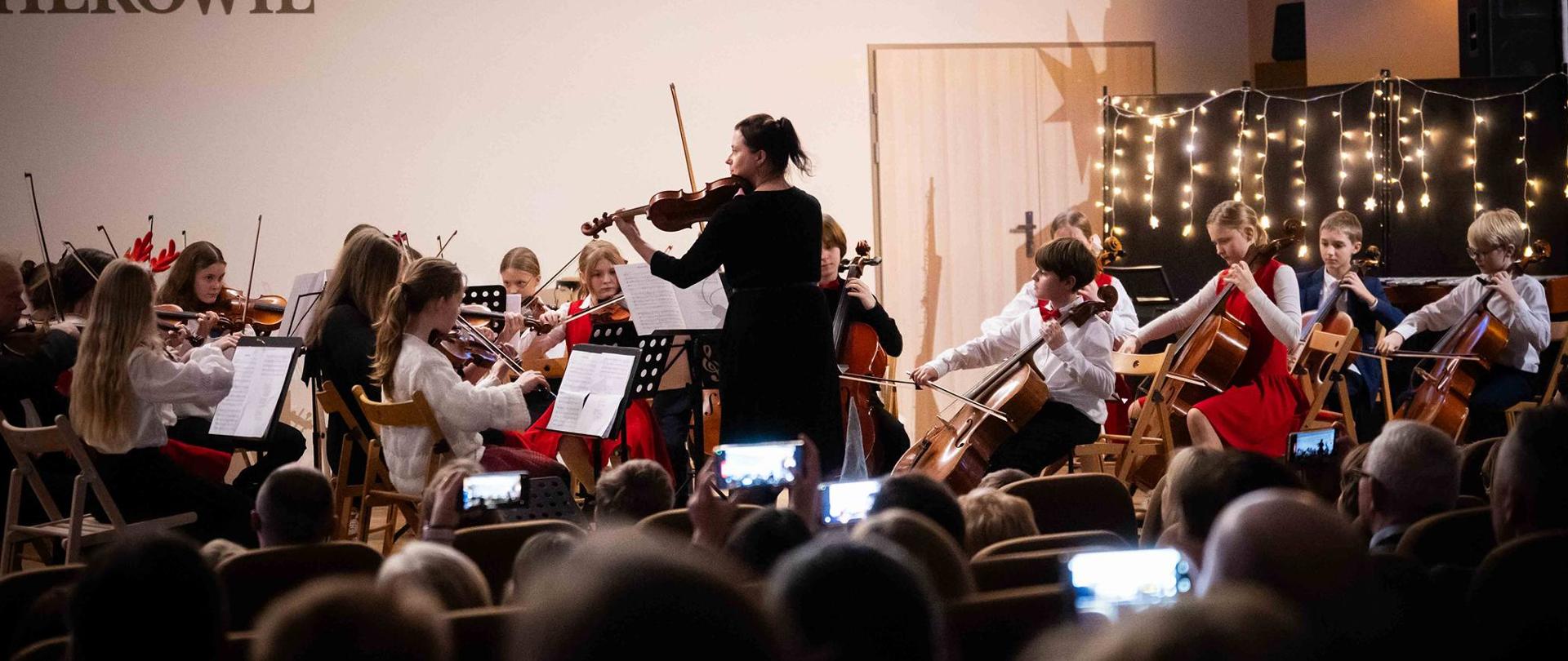 Orkiestra smyczkowa podczas koncertu kolęd. Tyłem stoi pani Agata Parzych, prowadząca orkiestrę