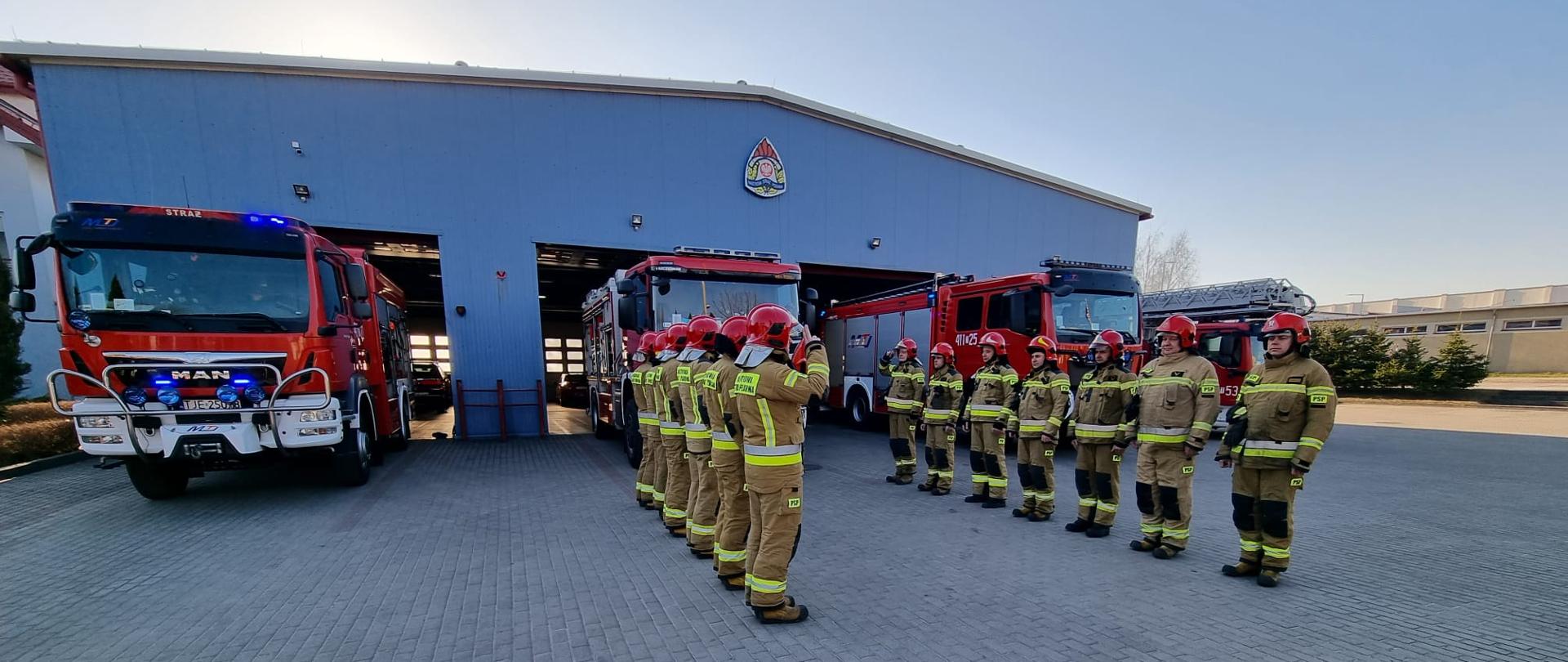Zdjęcie przedstawia 4 samochody specjalne Państwowej Straży Pożarnej na tle garażu jednostki ratowniczo-gaśniczej w Jędrzejowie. Przed pojazdami znajdują się strażacy w umundurowaniu specjalnym z założonymi czerwonymi hełmami na głowie. Strażacy oddają hołd ukraińskim strażakom, którzy stracili życie podczas służby ratując ludność cywilną w trakcie działań wojennych.