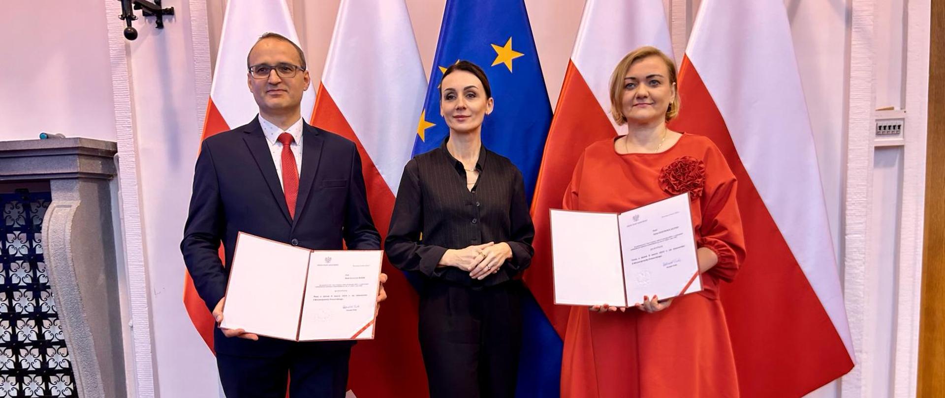 dwie kobiety i mężczyzna stoją w dużej sali na tle flag polskich i unijnej. trzymają w dłoniach teczki z nominacjami na wicewojewodów