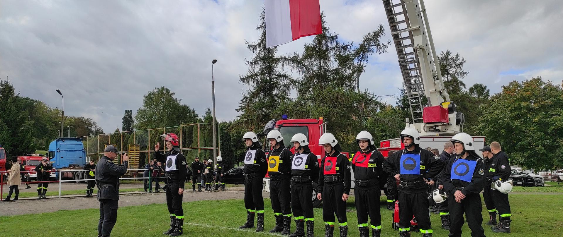 Na zdjęciu drużyna gotowa do ćwiczeń w mundurach oraz znacznikach. Z tyłu stoi podnośnik strażacki z podniesioną flagą państwową.