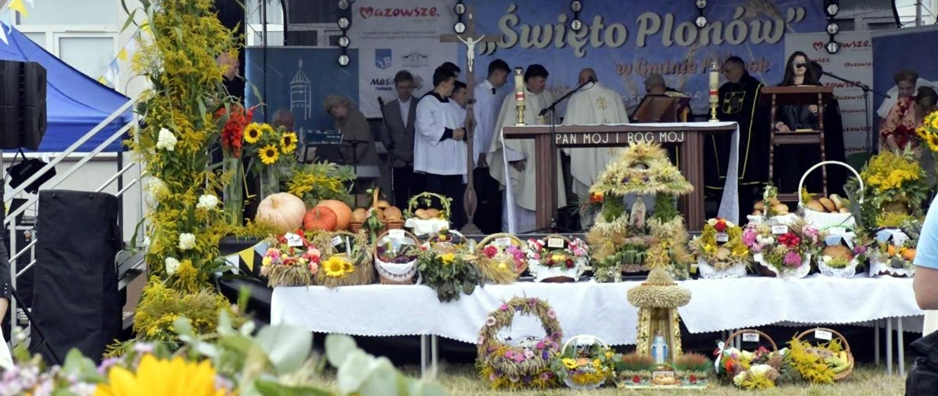 Na scenie podczas dożynek ustawione są stoły udekorowane wieńcami dożynkowymi, warzywami, owocami i kwiatami. W tle księża i uczestnicy uroczystości biorą udział w nabożeństwie pod napisem „Święto Plonów”.