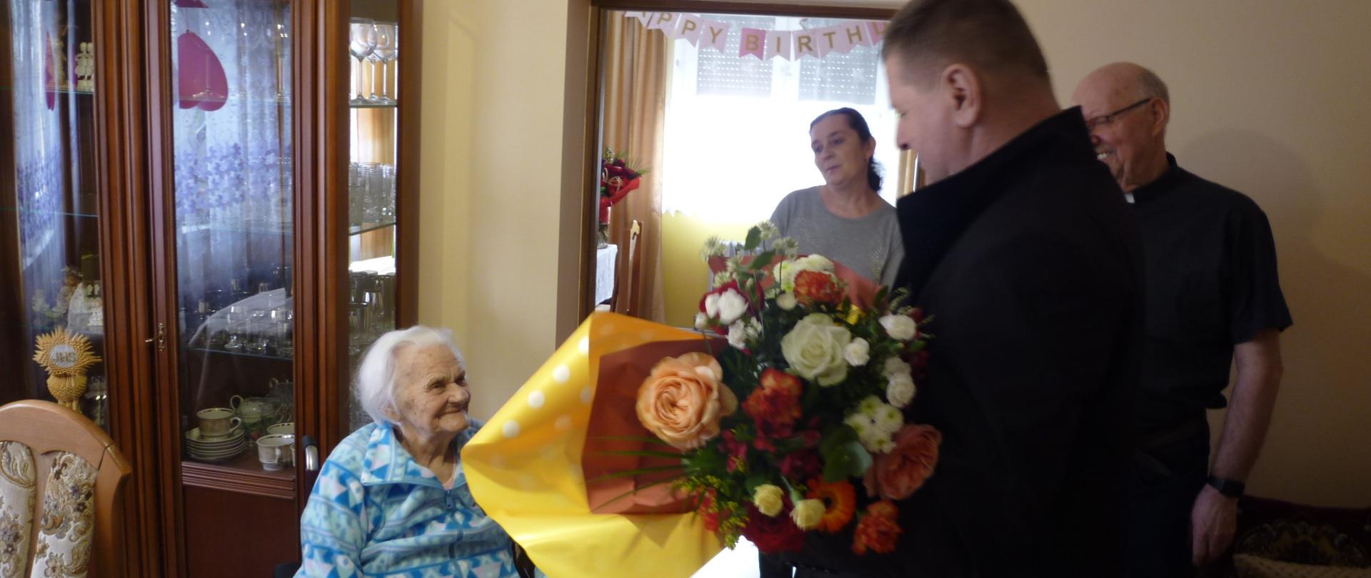 W pomieszczeniu znajduje się grupa osób. Od lewej: Jubilatka Pani Helena siedząca na wózku inwalidzkim, kolejno Pan Bogusław Gilarski Kierownik PT KRUS w Jarosławiu wręcza bukiet kwiatów Jubilatce. W oddali sytuacji tej przygląda się rodzina Jubilatki.