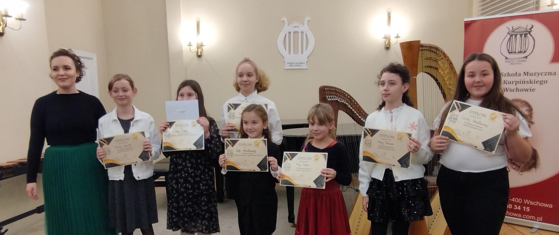 Uczestniczki konkursu harfowego z nauczycielką panią Dominiką Stachowicz w auli szkoły. Uczennice trzymają w dłoniach dyplomy.