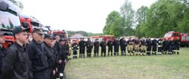 Strażacy PSP oraz OSP stoją w szeregu w ubraniach koszarowych na trawiastym placu OSP Marcinków w tle samochody bojowe oraz karetka pogotowia. 