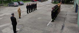 Widok z boku. Uroczysta zmiana służby w Jednostce Ratowniczo-Gaśniczej w Wadowicach. W szpalerze stoją zmiana przyjmująca i zdająca służbę. Dowódca czyta rozkaz dzienny. 