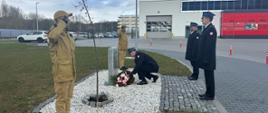 Komendant miejski składa kwiaty pod pomnikiem, za nim stoi delegacja funkcjonariuszy w mundurach wyjściowych. Obok pomnika stoi dwóch strażaków.