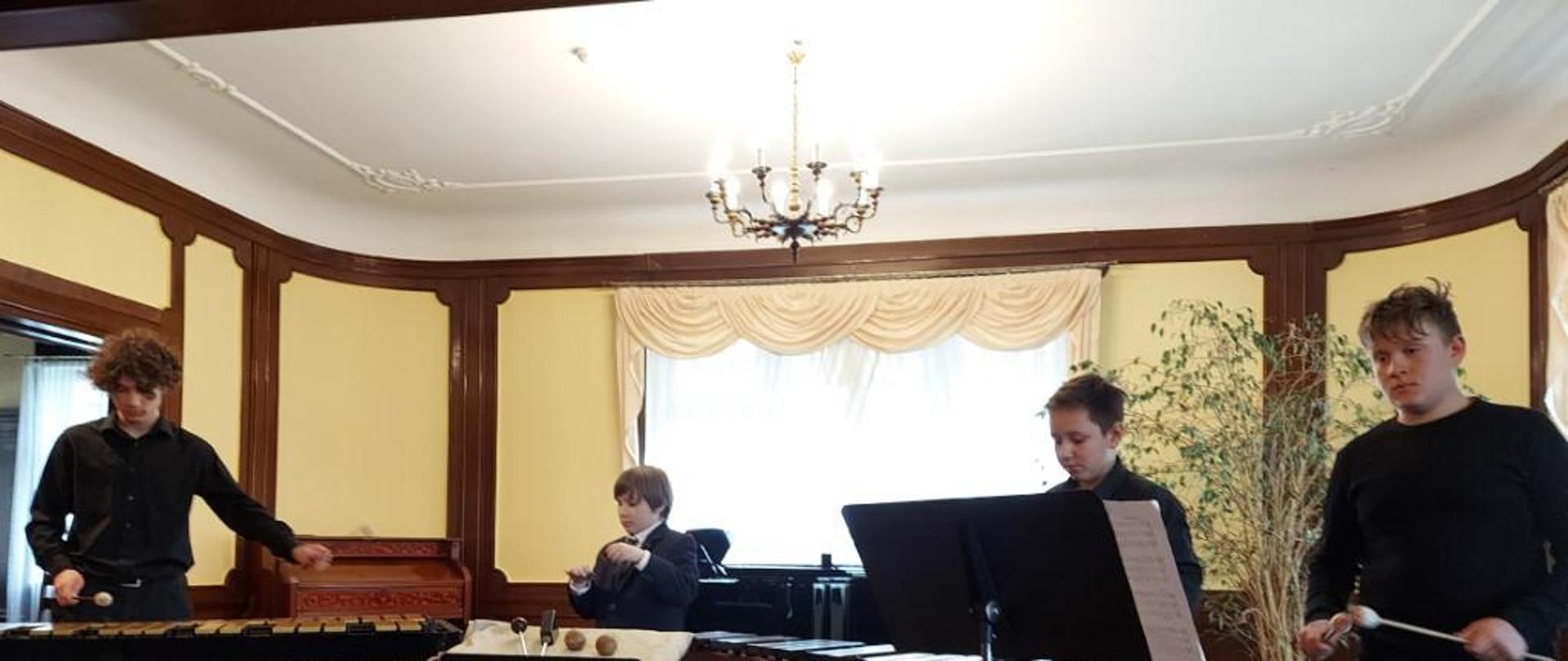 Zdjęcie kolorowe. Czterech chłopców gra na instrumentach perkusyjnych. W tle czarny fortepian i zabytkowa brązowa fisharmonia.