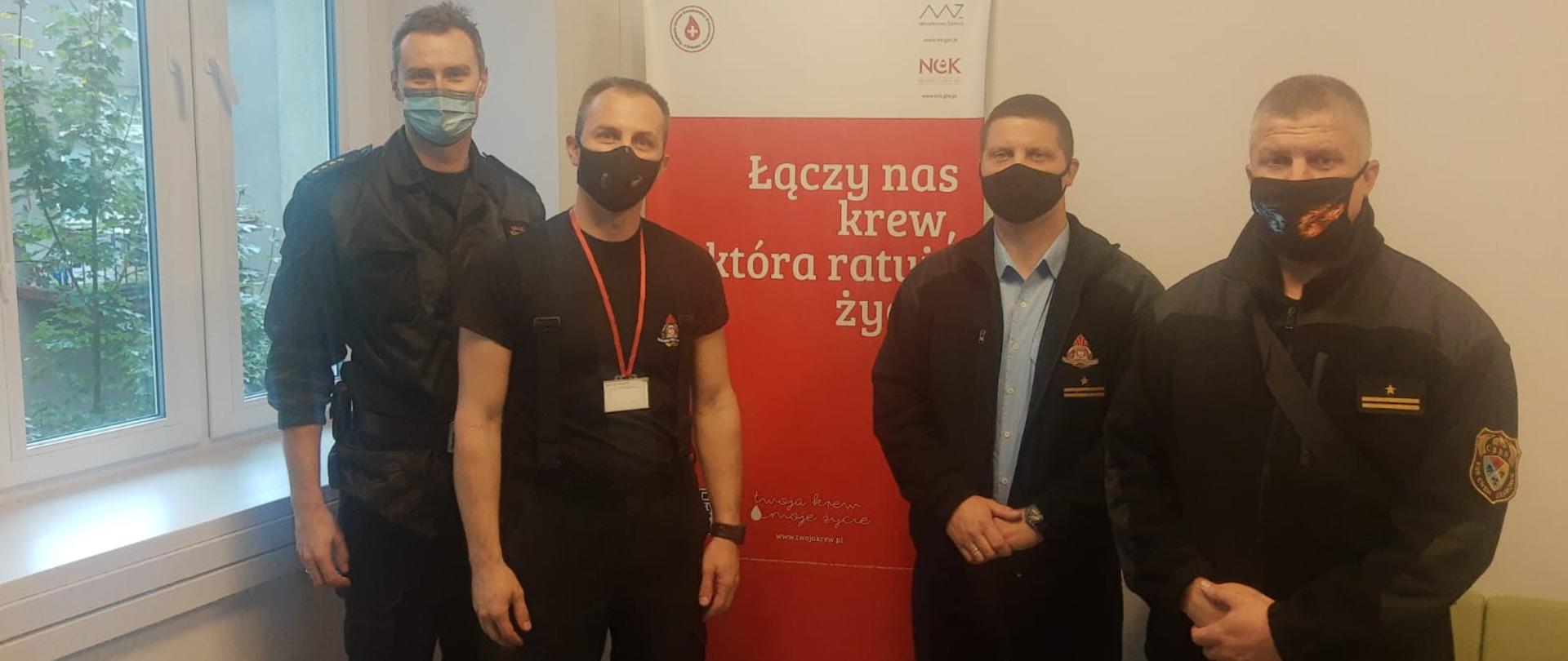 Zdjęcie przedstawia czterech strażaków Komendy Miejskiej PSP w Krakowie w maseczkach na twarzach na tle baneru "Łączy nas krew, która ratuje życie".