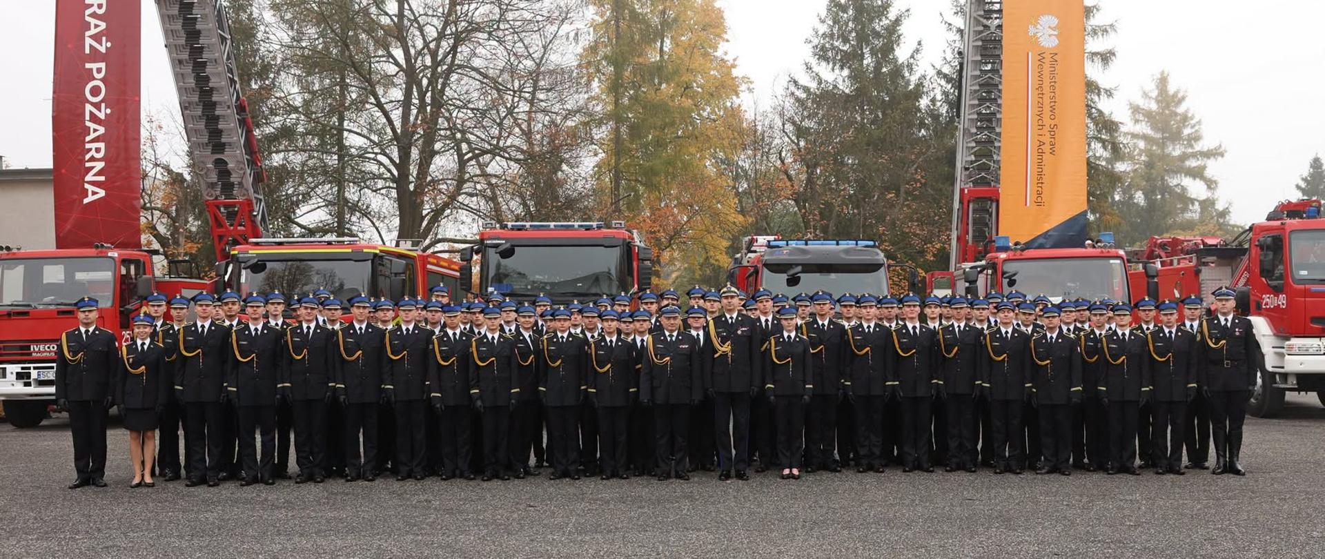8czestochowa.jpeg – Pamiątkowe zdjęcie dużej grupy kadetów w galowych mundurach ustawionych przed samochodami pożarniczymi. Po obu stronach wzniesione są drabiny mechaniczne z pionowymi banerami Państwowej Straży Pożarnej oraz ochrony ludności i obrony cywilnej.
