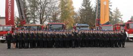 8czestochowa.jpeg – Pamiątkowe zdjęcie dużej grupy kadetów w galowych mundurach ustawionych przed samochodami pożarniczymi. Po obu stronach wzniesione są drabiny mechaniczne z pionowymi banerami Państwowej Straży Pożarnej oraz ochrony ludności i obrony cywilnej.
