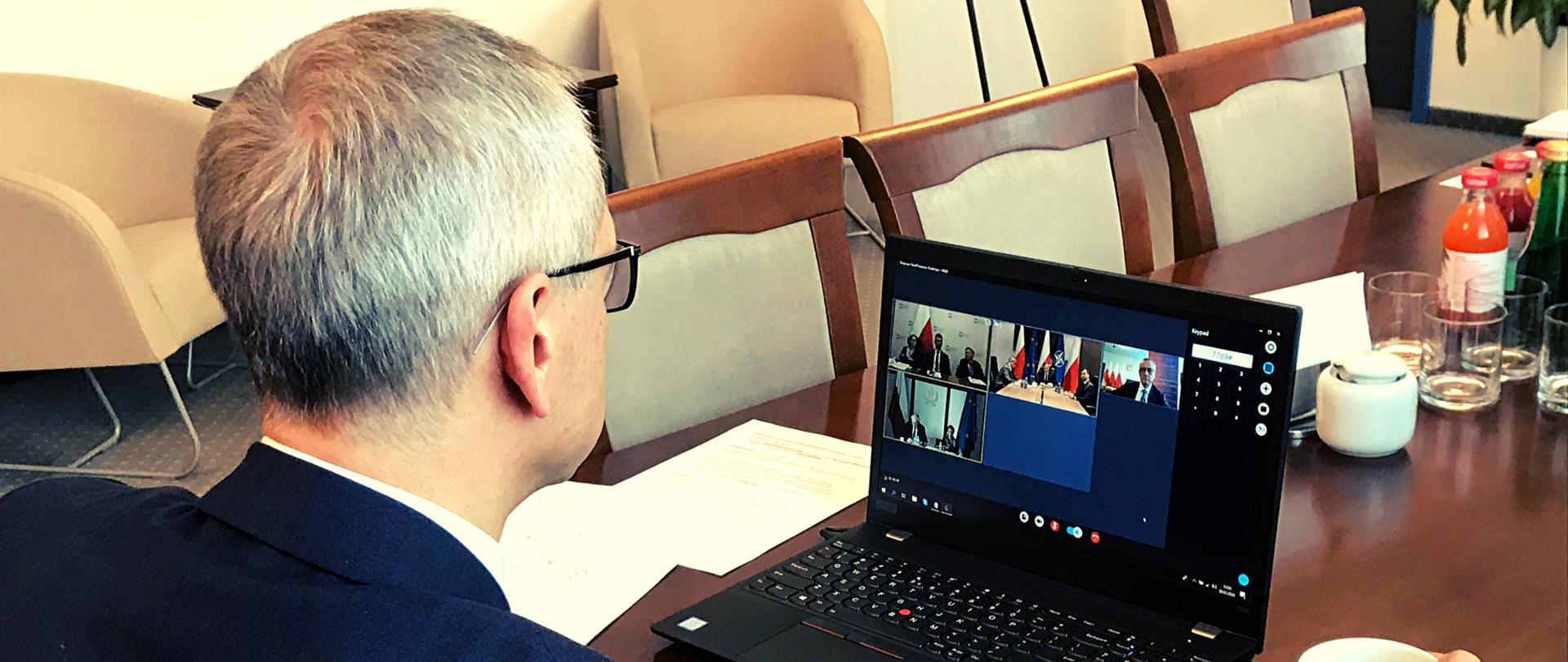 Marek Zagórski w gabinecie. Siedzi przy stole, na którym stoi laptop. Na ekranie widać okienka z wizerunkami osób biorących udział w wideokonferencji. Ujęcie z zza ramienia ministra.