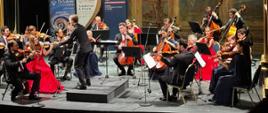 Orkiestra kameralna na scenie pod batutą Christopha Koncza. Kobiety w czerwonych sukniach, mężczyźni w czarnych garniturach, białych koszulach i czerwonych krawatach. W tle roll-upy Ambasady RP w Valletcie i De Vallette Chamber Orchestra.