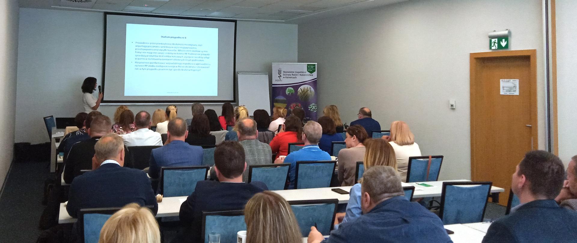 Na zdjęciu widać uczestników narady słuchających wykładowcy i oglądających prezentację wyświetlaną na rzutniku.