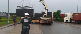 Na pierwszym planie umundurowany inspektor ITD. W tle stoi samochód ciężarowy z żurawiem i przeładowuje nadwyżkę szamb betonowych na podstawioną przyczepę ciężarową.
