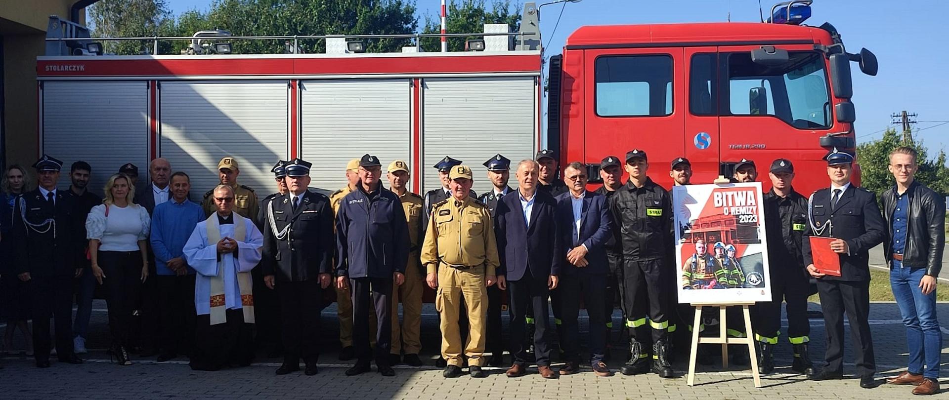 Fotografia wykonana podczas uroczystości związanej z przekazaniem samochodu pożarniczego dla OSP Grochowe I. Na zdjęciu na tle przekazanego samochodu pożarniczego stoją zaproszeni na uroczystość goście. Na pierwszym planie zaprezentowano plakat reklamujący akcję "Bitwa o Remizy".
