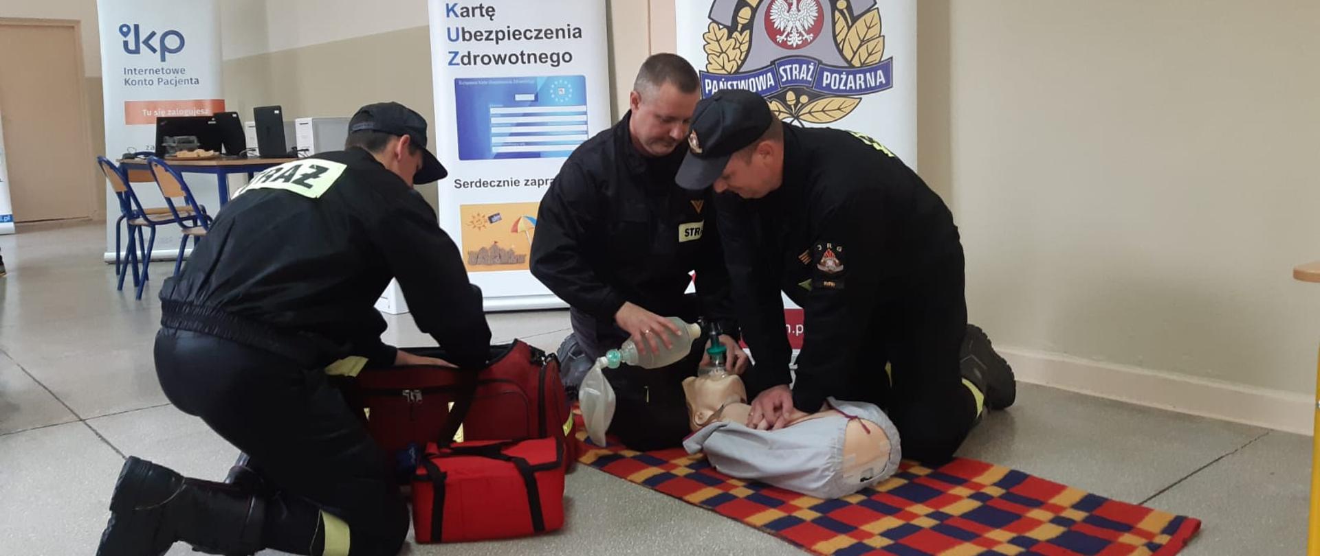 Trzech strażaków robi pokaz pierwszej pomocy na fantomie. Leży on na kocu w budynku. W tle baner PSP oraz NFZ.
