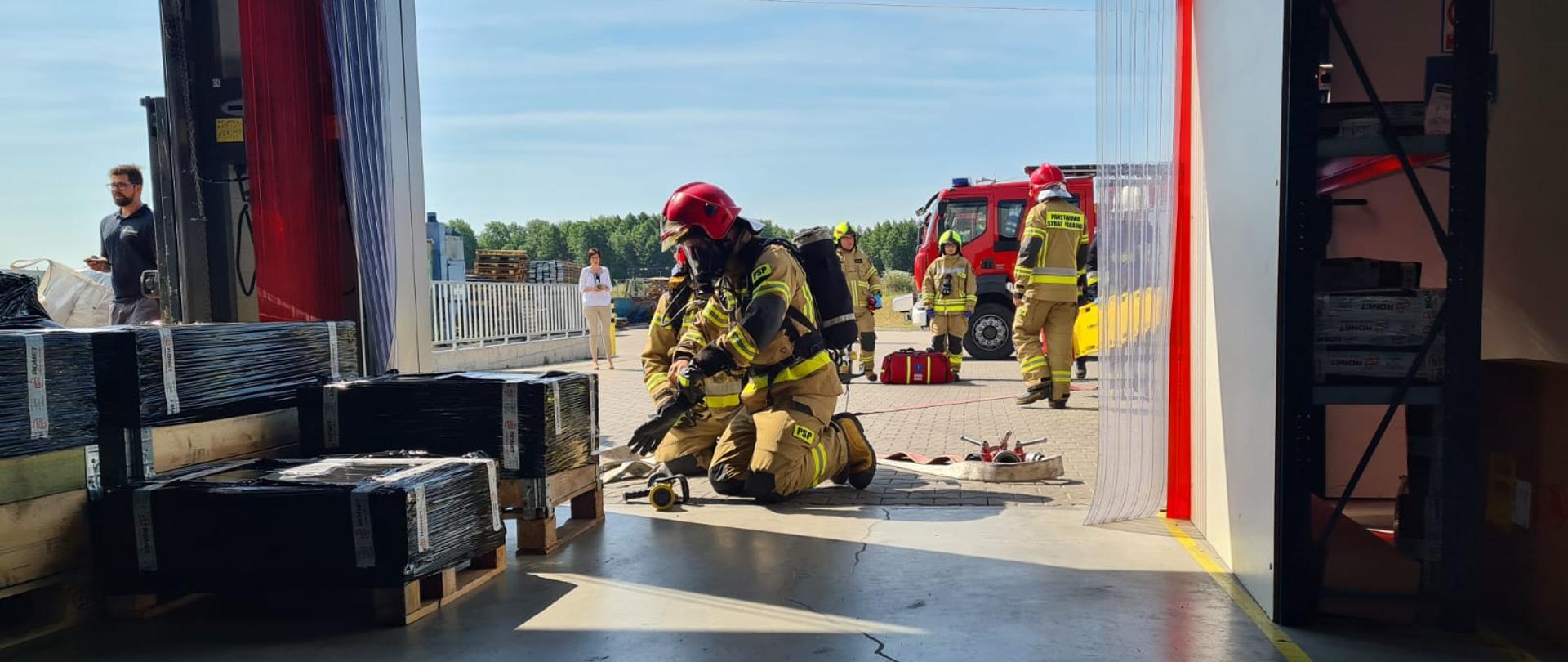 Zdjęcie wykonano w magazynie firmy Ronet w ciągu dnia. Widok na bramę magazynową otwartą, po lewej stronie stoi kilka zafoliowanych palet. W bramie dwóch strażaków Państwowej Straży Pożarnej w Ubraniach specjalnych z aparatami ochrony układu oddechowego, jeden z nich ubiera rękawice specjalne obok nich leży linia wężowa z prądownicą po lewej stronie oraz rozdzielaczem po prawej stronie. W tle na parkingu widocznych trzech strażaków Ochotniczej Straży Pożarnej oraz jeden strażak Państwowej Straży Pożarnej, za nimi stoi średni samochód gaśniczy. 