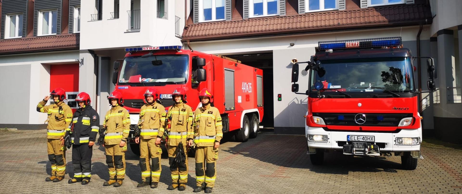 Na zdjęciu widać sześciu strażaków stojących w jednym szeregu oraz dwa samochody strażackie na placu przed budynkiem Komendy Powiatowej PSP w Łęczycy. Pięciu strażaków jest w żółtych ubraniach a jeden w czarnych. Wszyscy strażacy mają czerwone hełmy. Jest widno.