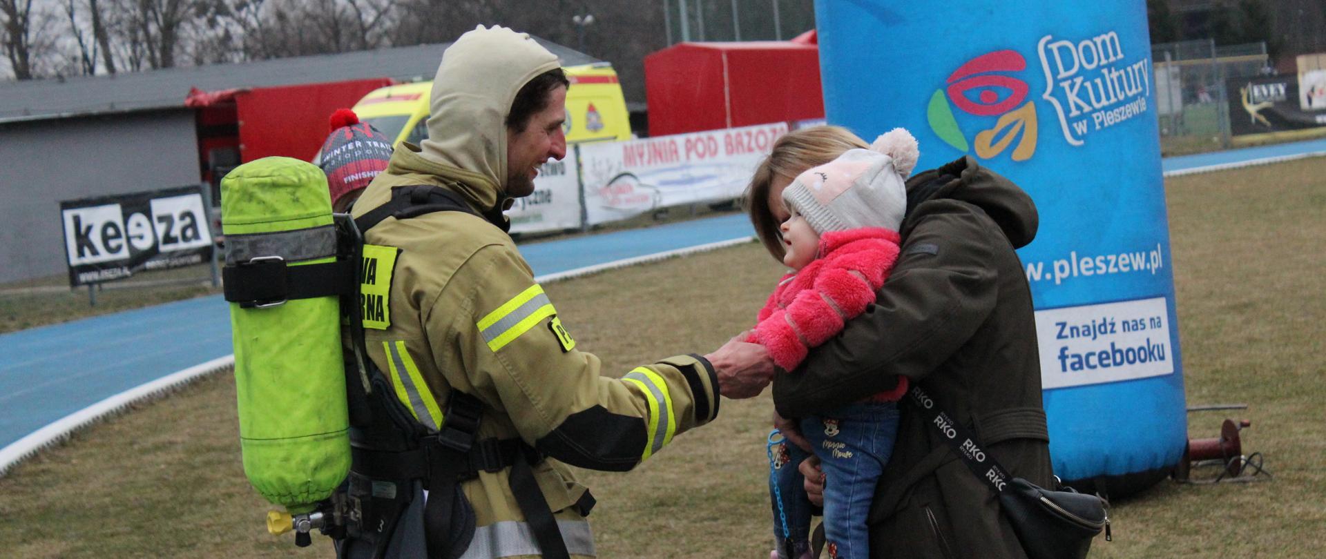 Strażak ubrany w ubranie specjalne z butlą aparatu oddechowego na plecach podaje ręke malutkiej dziewczynce - Jagódce-, któr znajduje się na ekach jej mamy. w tle widoczny tor i część bramy startowej biegu