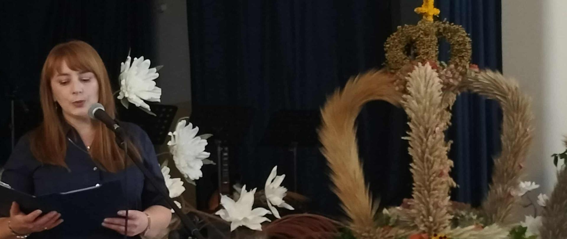 Po lewej stronie na pierwszym planie stoi słuchaczka Studium, która przemawia do mikrofonu. Tuż za nią są białe kwiaty oraz stół na którym stoi wieniec dożynkowy, który zrobiony jest z kłosów zbóż oraz kwiatów. Ma on kształt korony. Na samej górze jest plecionka w kształcie serca oraz pleciony krzyż. 