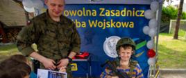 Dziecko ubrane w strój wojskowy uczestniczy w zajęciach edukacyjnych, stojąc w pobliżu stanowiska tematycznego z elementami wyposażenia wojskowego.