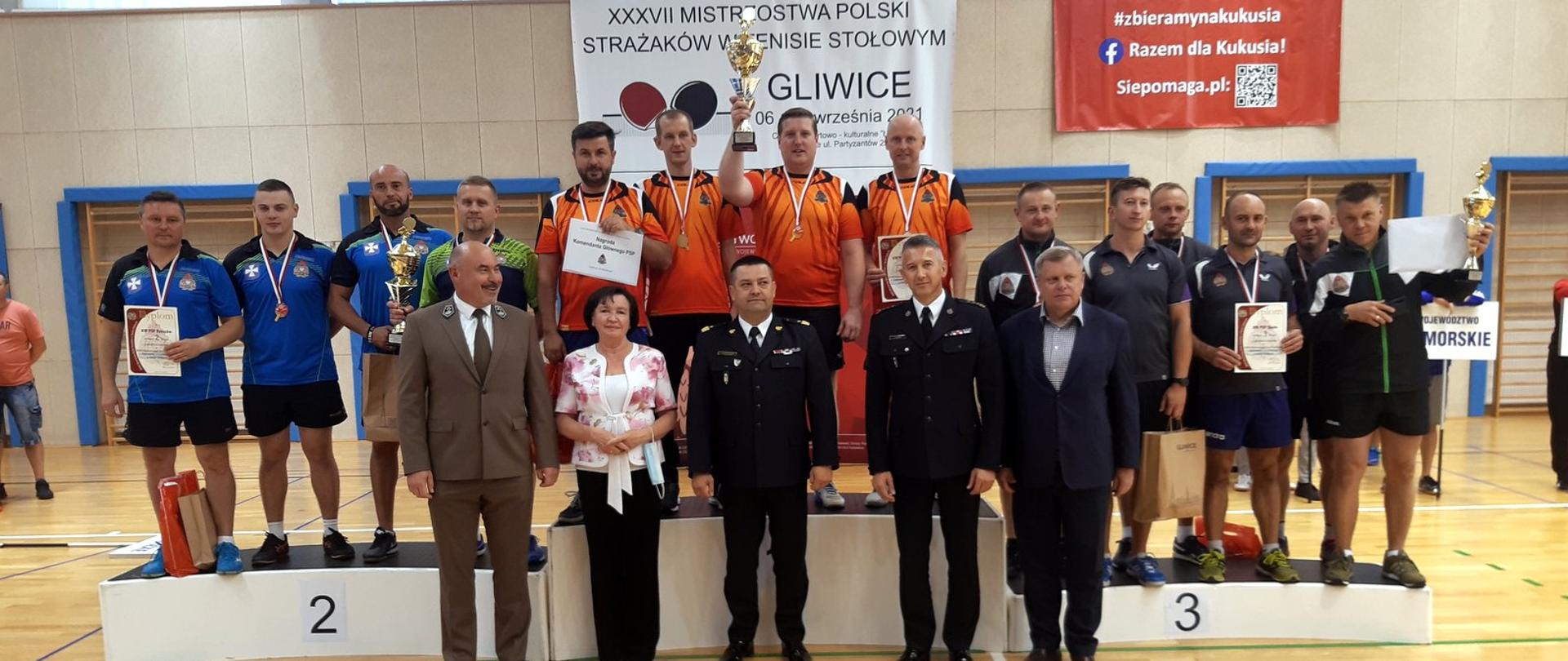Podium ustawione na sali sportowej. Na stopniach podium stoi grupa zawodników z pucharami, medalami i dyplomami. Przed podium ustawionych jest pięć osób, wśród nich dwóch mężczyzn w wyjściowych mundurach PSP.