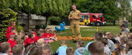 Zdjęcie przedstawia grupę przedszkolaków słuchającą strażaka. Na drugim planie znajduje się samochód pożarniczy.