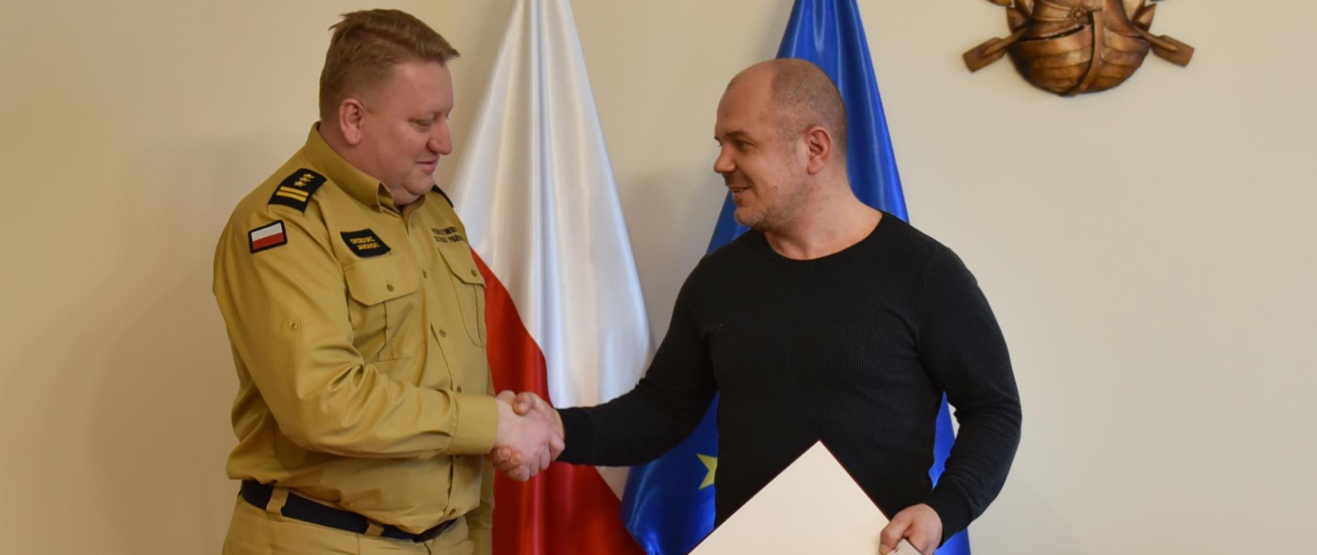 na zdjęciu strażak w umundurowaniu służbowym w kolorze piaskowym oraz mężczyzna w swetrze i jeansach, który trzyma w ręce teczkę z dokumentem, obaj uściskają sobie dłonie w tle flagi polski i unii europejskiej,
