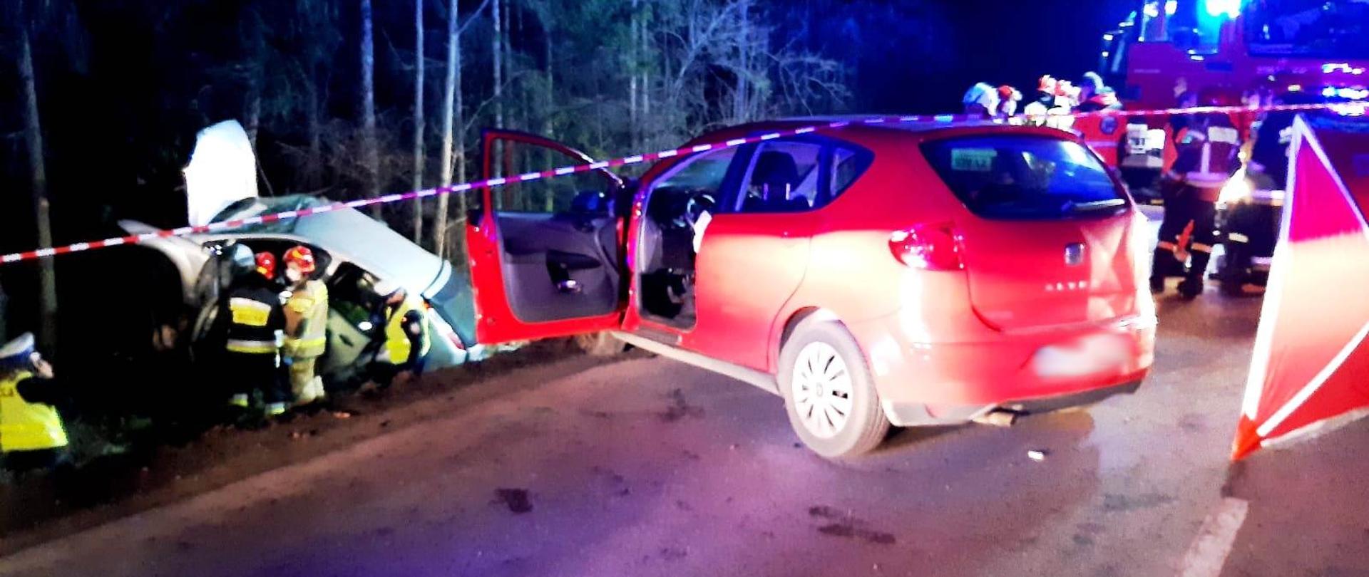 Dwa rozbite samochody. jeden jasnego koloru poza drogą. Obok niego strażacy. Drugi czerwony stoi w poprzek drogi na jednym pasie. Obok niego stoją również strażacy. Rozłożony parawan ochronny. W tle samochody pożarnicze.