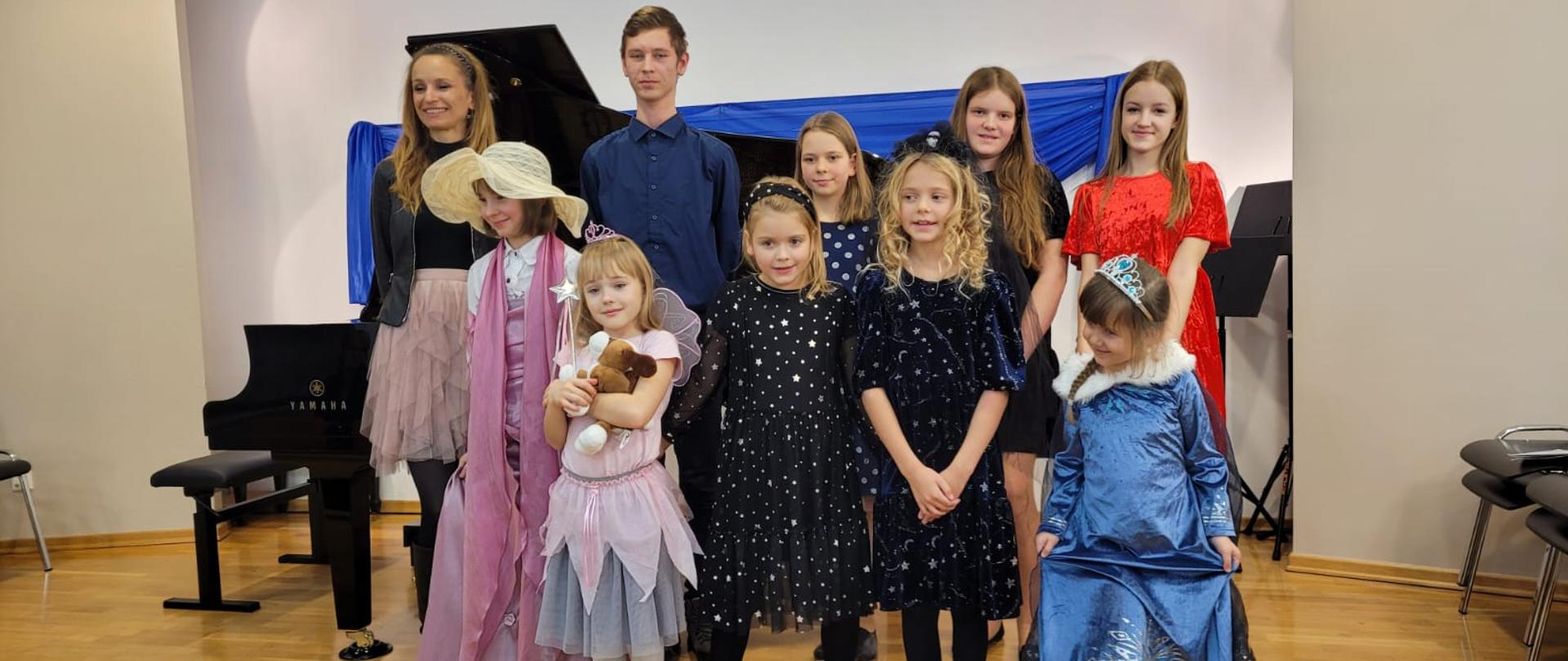 Na zdjęciu uczniowie klasy fortepianu na scenie. Niektóre dzieci w zabawnych przebraniach: wróżki, księżniczki. W tle czarny fortepian.