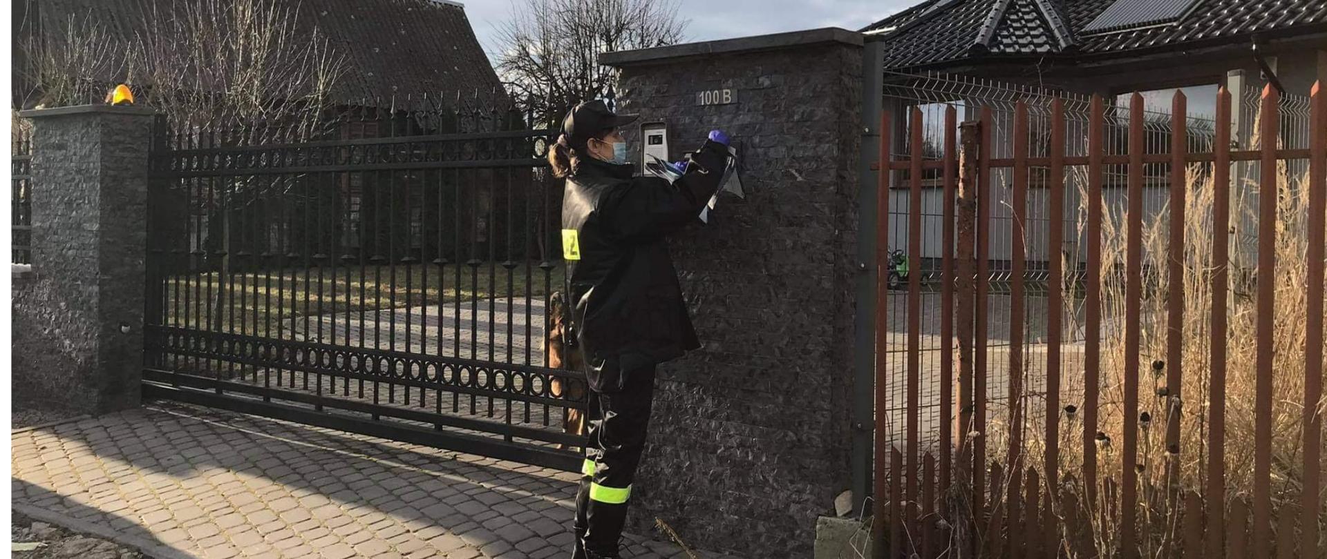 Fotografia przedstawiająca strażaka OSP rozwieszającego ulotki informacyjne na ogrodzeniu na dotyczące szczepień przeciwko COVID-19