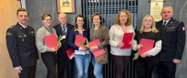 Grupa ośmiu osób pozuje do zdjęcia w eleganckim wnętrzu z ciemnym, nowoczesnym wystrojem. Dwie osoby w czarnych mundurach straży pożarnej stoją po bokach grupy, a między nimi znajdują się cywilni pracownicy PSP – pięć kobiet i jeden mężczyzna. Każda z osób trzyma czerwoną teczkę z godłem Rzeczypospolitej Polskiej, a niektóre również kwiaty – pojedyncze tulipany.