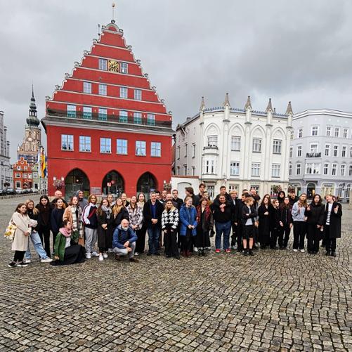 zdjęcie przedstawia uczestników warsztatów na środku rynku zabytkowej miejscowości, w tle kamienice i ratusz