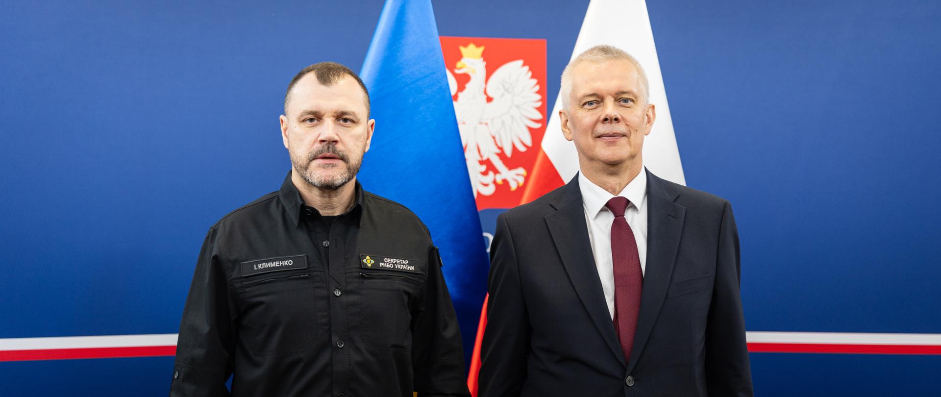 Na zdjęciu minister Tomasz Siemoniak (po prawej) oraz Ihor Klymenko, sekretarz Rady Bezpieczeństwa Narodowego i Obrony Ukrainy (po lewej). Za ministrami stoją dwie flagi – po prawej stronie biało-czerwona flaga Polski oraz po lewej niebiesko-żółta flaga Ukrainy. Pomiędzy flagami godło Polski – biały orzeł na czerwonym tle. W tle niebieska ścianka z biało-czerwonym paskiem na dole zdjęcia.