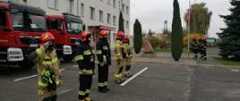 Strażacy stoją na baczność na placu przed budynkiem komendy, w tle samochody pożarnicze.