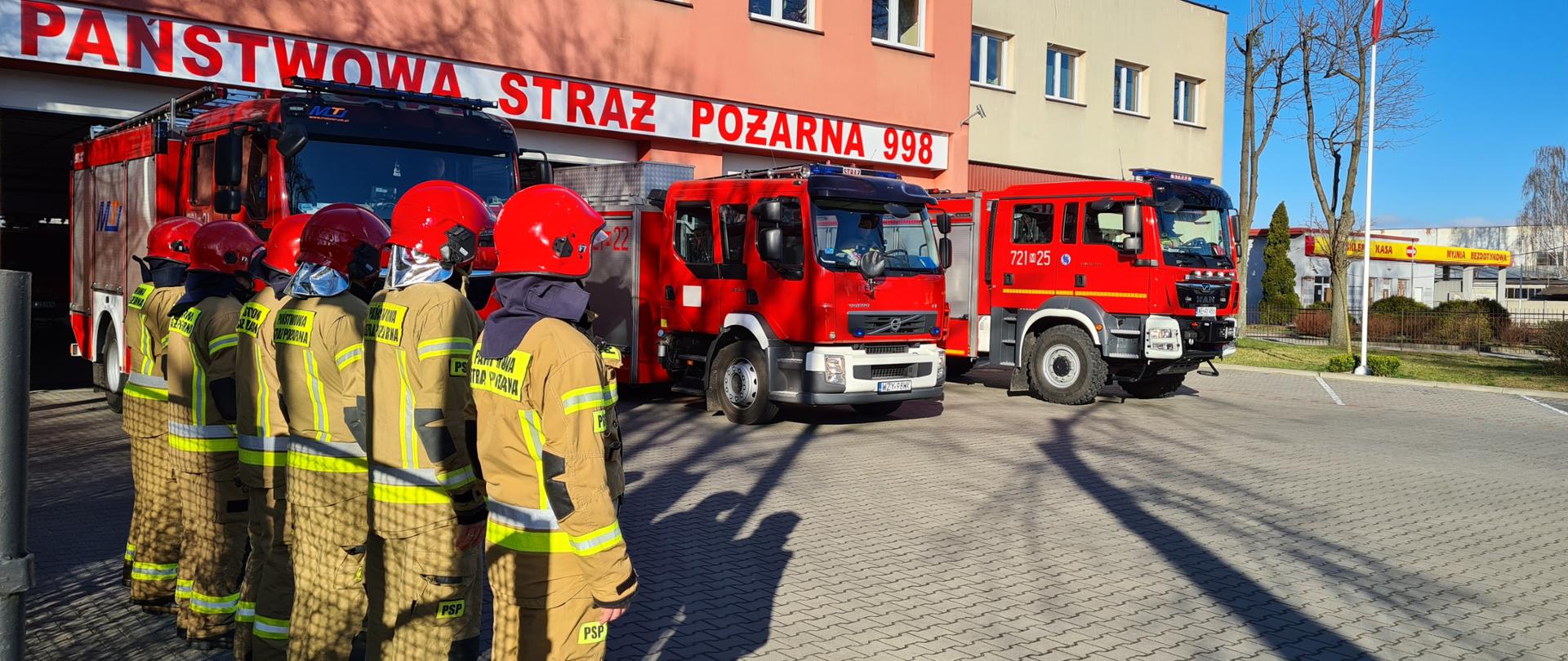 Na placu zewnętrznym przed komendą stoją wystawione przed garaże samochody bojowe. Przed pierwszym z nich w szeregu stoją strażacy w mundurach bojowych.