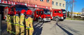 Na placu zewnętrznym przed komendą stoją wystawione przed garaże samochody bojowe. Przed pierwszym z nich w szeregu stoją strażacy w mundurach bojowych.