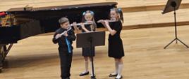 Dwie dziewczynki i chłopiec stoją na środku estrady sali koncertowej PSM przy pulpicie i grają na fletach poprzecznych. Za nimi stoi fortepian.