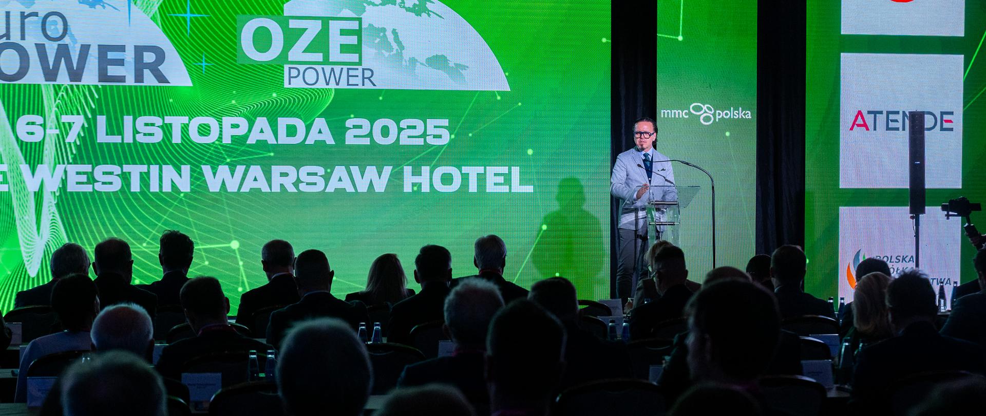 Minister Wojciech Balczun stoi za mównicą. W tle ekran. Ujęcie zza słuchaczy. 