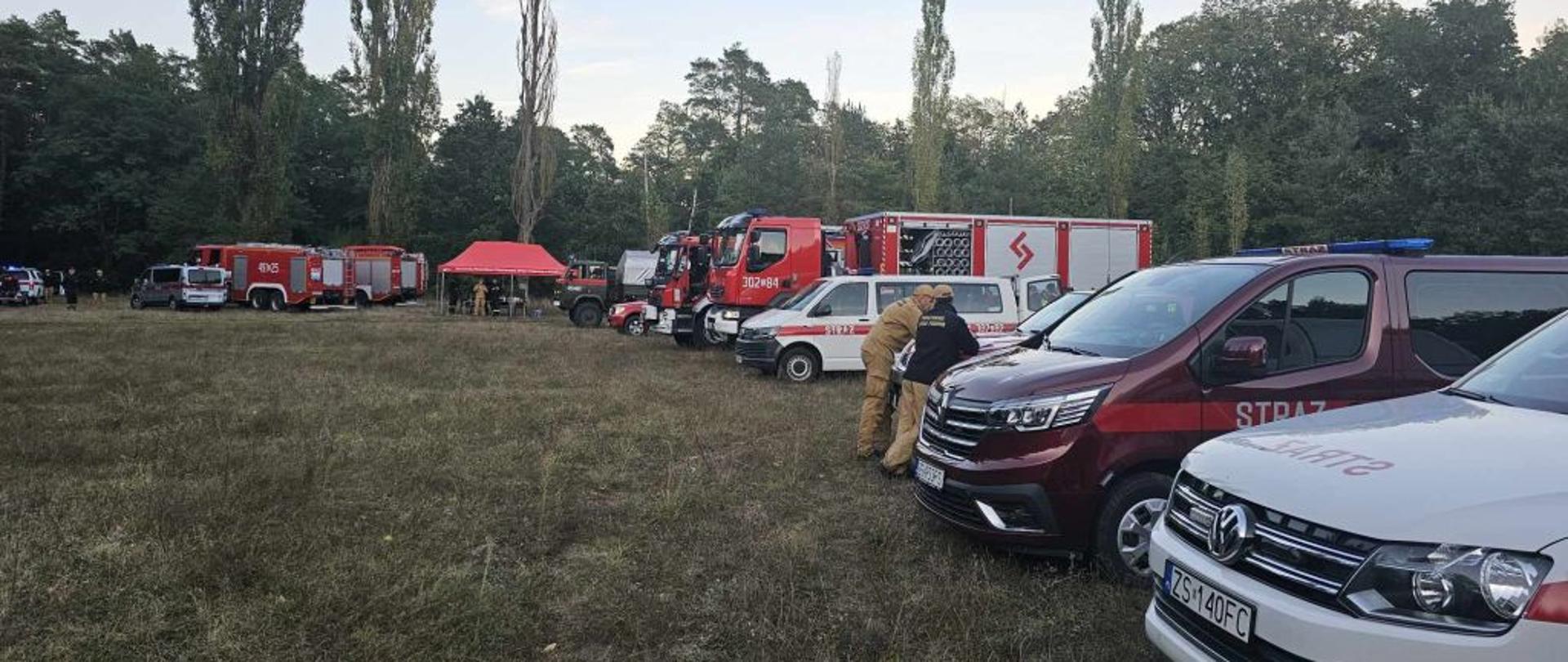 Zdjęcie wykonane w dzień. Na kawałku łąki, placu stoją wozy strażackie. W tle widać wysokie drzewa. Przy jednym z wozów grupka strażaków.