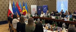 Uczestnicy programu i jego organizatorzy podczas spotkania podsumowującego program - w reprezentacyjnej sali w Kancelarii Prezesa Rady Ministrów kilkanaście osób siedzi przy owalnym stole, w tle flagi państw uczestniczących w programie
