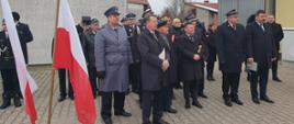 Zdjęcie przedstawia strażaków, ministra, posła, radnego oraz inne osoby stojące na placu przez remizą OSP podczas uroczystej zbiórki 