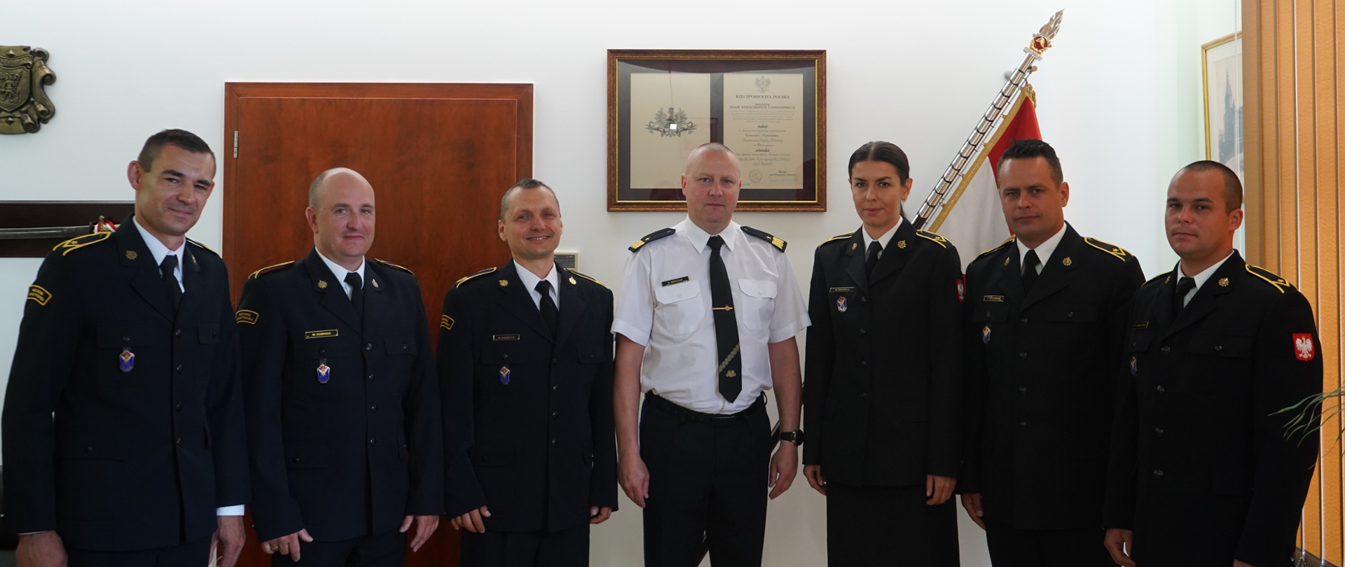 grupa strażaków w ciemnych mundurach z białymi koszula i krawatem stoi obok siebie, w środku stoi komendnt w białej koszuli z krawatem i pagonami. w tle sztandar