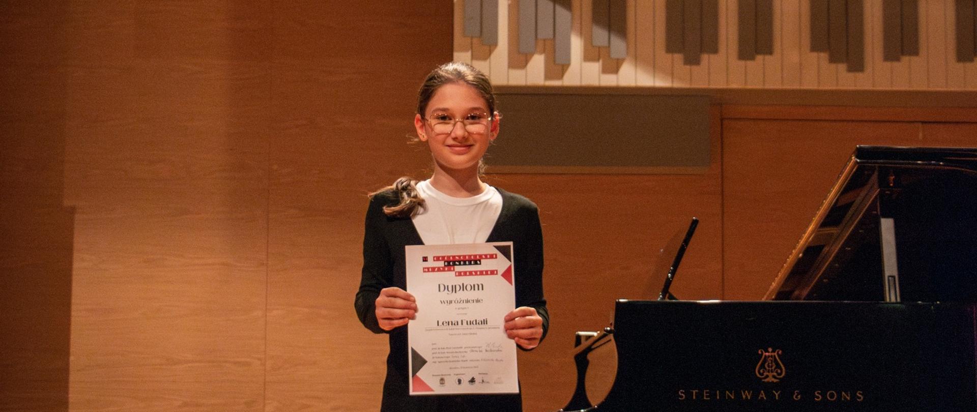 Młoda osoba z długimi, ciemnymi włosami spiętymi z tyłu, w okularach, czarno-białej bluzce i czarnych spodniach, stoi na drewnianej scenie. Trzyma w obu rękach dyplom skierowany w stronę widza. Na dyplomie widoczne są polskie napisy, w tym słowo "Dyplom" i "I miejsca".
Za nią, po prawej stronie, stoi czarny fortepian Steinway & Sons z otwartą klapą. W tle znajduje się ściana z drewnianą boazerią, a nad nią fragmenty organów piszczałkowych. Po lewej stronie, nad głową osoby, wisi baner informujący o "Ogólnopolskim Konkursie Muzyki Polskiej" w Jarosławiu w 2025 roku, z elementami przypominającymi klawiaturę fortepianu i czerwonymi akcentami. Oświetlenie sceniczne jest skierowane na osobę trzymającą dyplom.