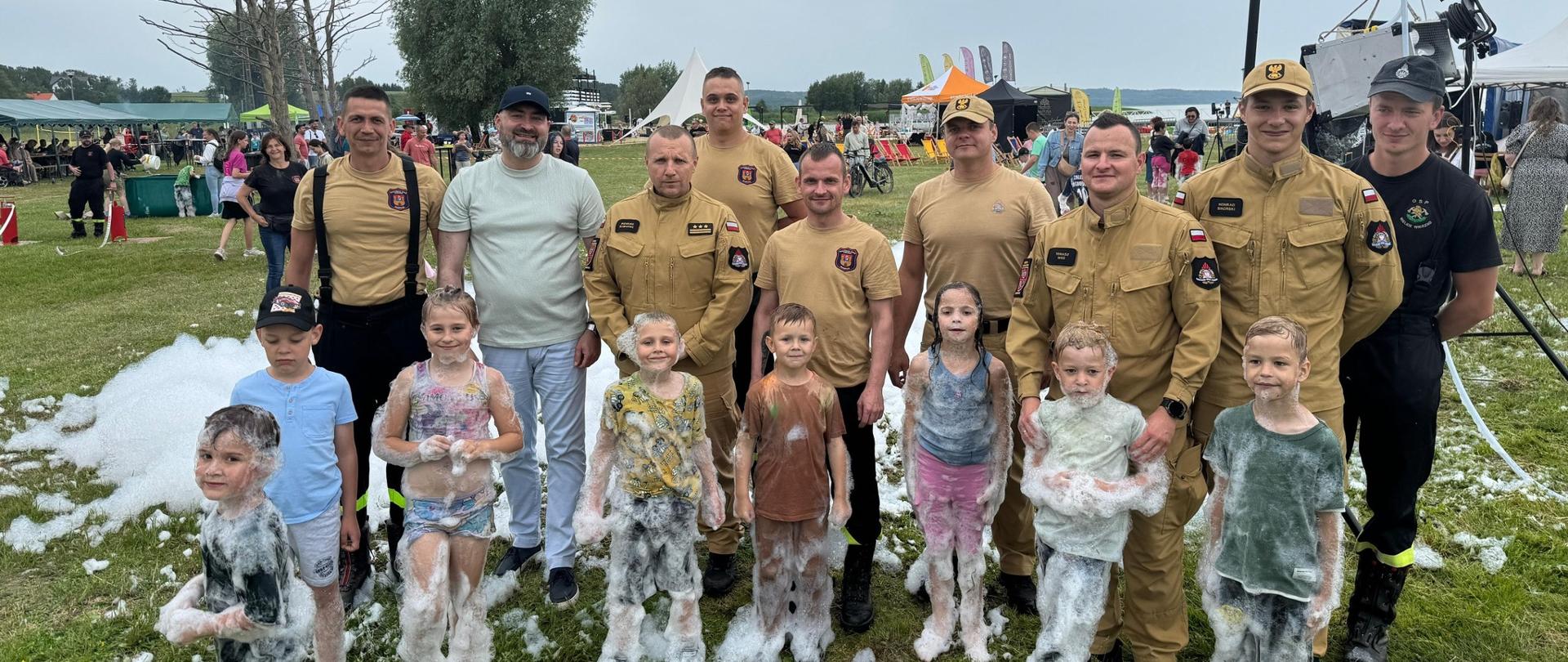 Zielona polana. Do zajęcia pozuje grupa strażaków w jasnych mundurach, przed nimi dzieci oblepione pianą. Pochmurnie.