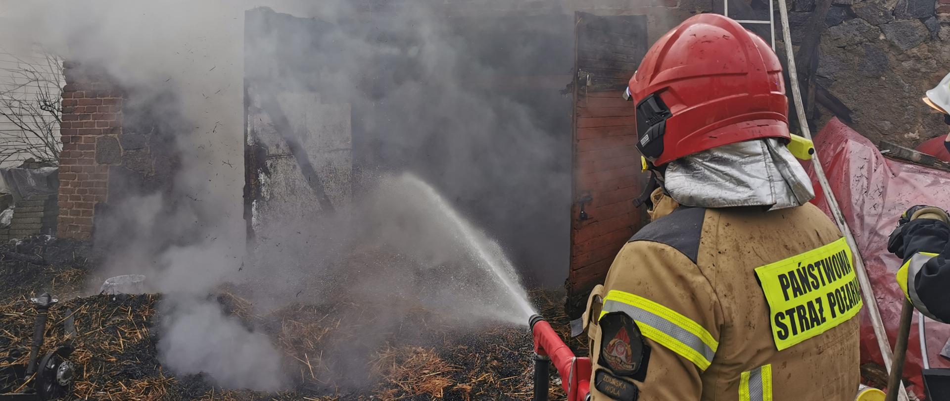 Zdjęcie przedstawia strażaka z Państwowej Straży Pożarnej gaszącego wodą słomę zrzuconą z poddasza budynku gospodarczego. W górnej części zdjęcia widać strażaków gaszących poddasze. 
