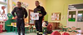 Na obrazie widzimy dwóch strażaków, mężczyzn znajdujących się w pomieszczeniu przedszkola. Wzrok strażaków skierowany jest przed siebie, dokonują przemówienia do grupy odbiorców którzy nie są widoczni. Strażacy ubrani są w czarne mundury sztabowe, wysokie specjalistyczne buty koloru czarnego, pasy na biodrach. Strażak z prawej strony kadru trzyma kartkę na której widzimy namalowany wóz strażacki i strażaków gaszących pożar. Akcja ma miejsce w pomieszczeniu przedszkola zielone ściany, mebelki na których znajdują się pomoce dydaktyczne zielona podłoga ( wykładzina ), małe stoliki i krzesełka z zielonymi nogami, dwa czerwone hełmy oraz ubranie specjalne leżą na stoliku. Za strażakami widzimy białe przeszklone drzwi wejściowe. 