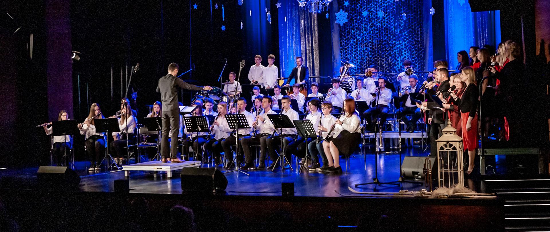 Na zdjęciu, w sali widowiskowej Jasielskiego Domu Kultury widnieje Młodzieżowa Orkiestra Dęta pod dyrekcją Piotra Dudy oraz Zespół Wokalny Jasielskiego Domu Kultury. Kolorystyka zdjęcia jest niebiesko-czarna.